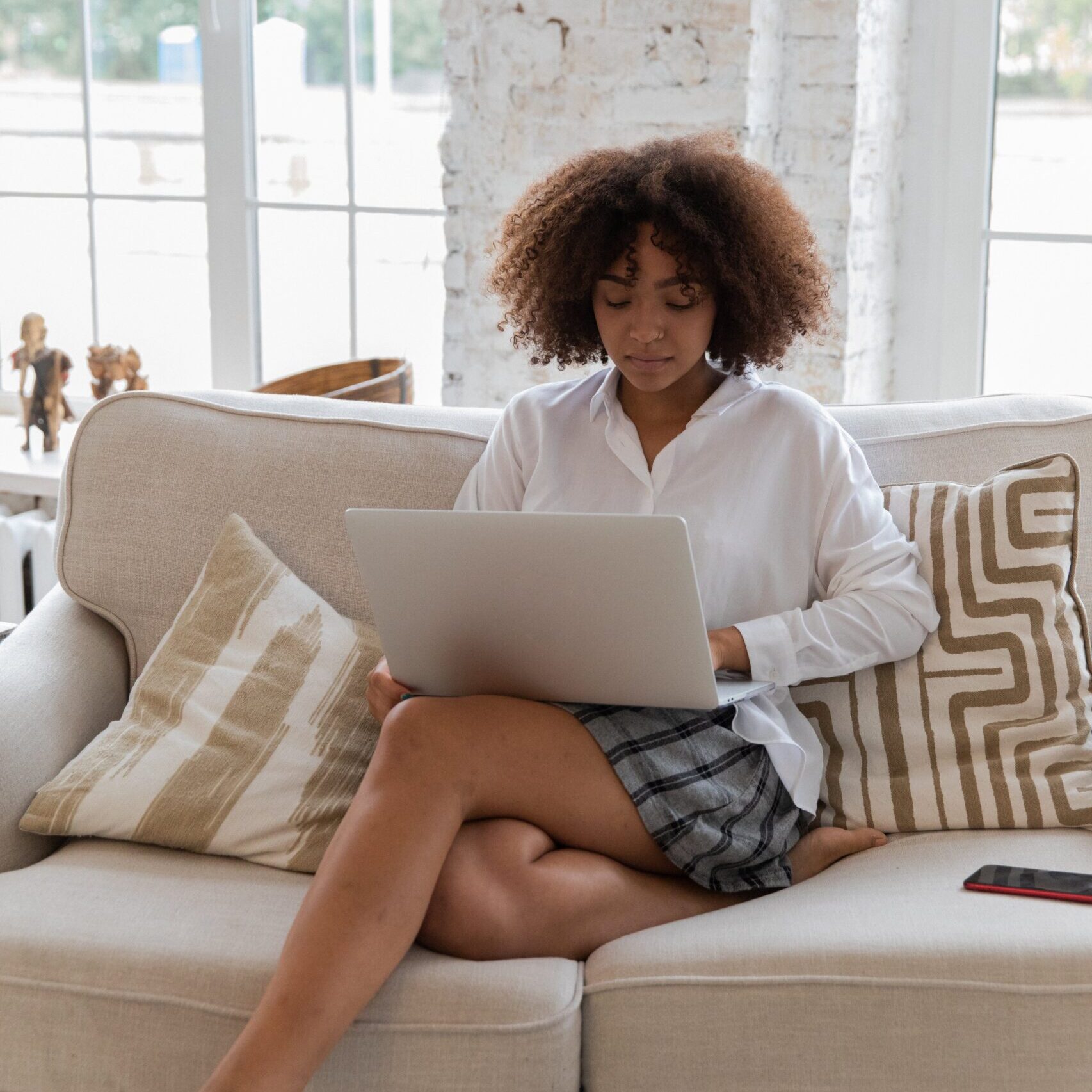 Black woman with brown curly hair in a white blouse and blue/white striped shorts sitting crossed legged on a cream couch using her laptop